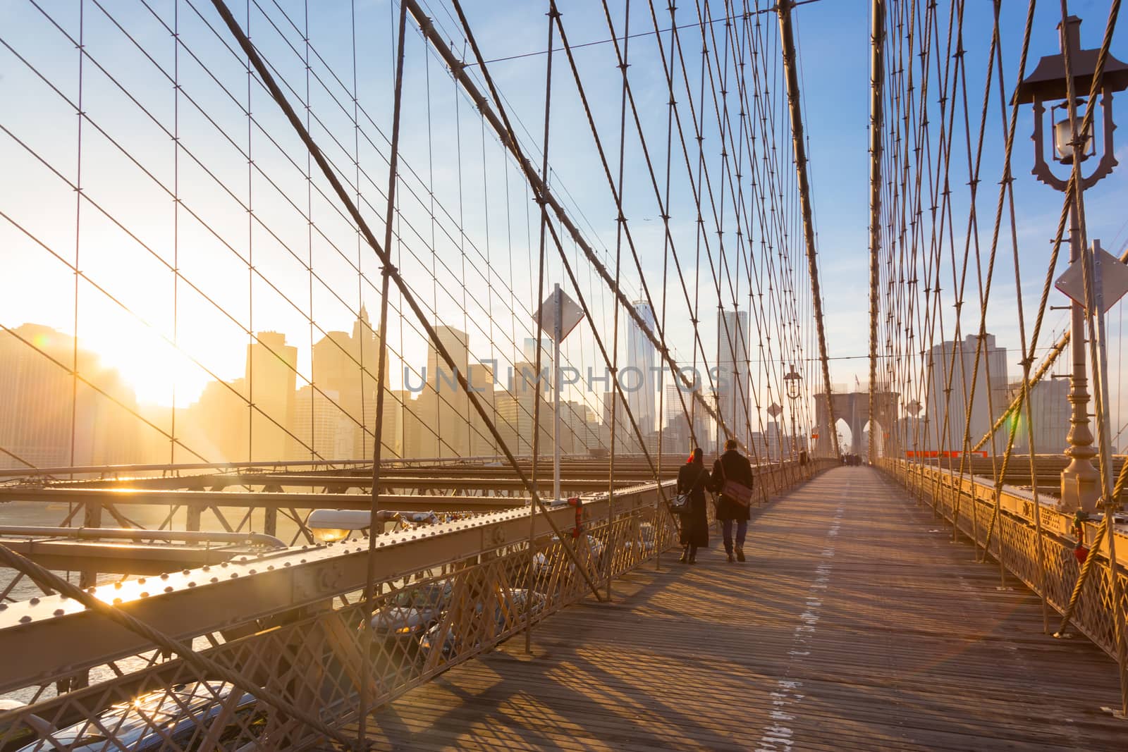 Couple walking on pedestrian path across Brooklyn bridge. New York City Manhattan downtown skyline in sunset with skyscrapers illuminated over East River panorama as seen from Brooklyn bridge.