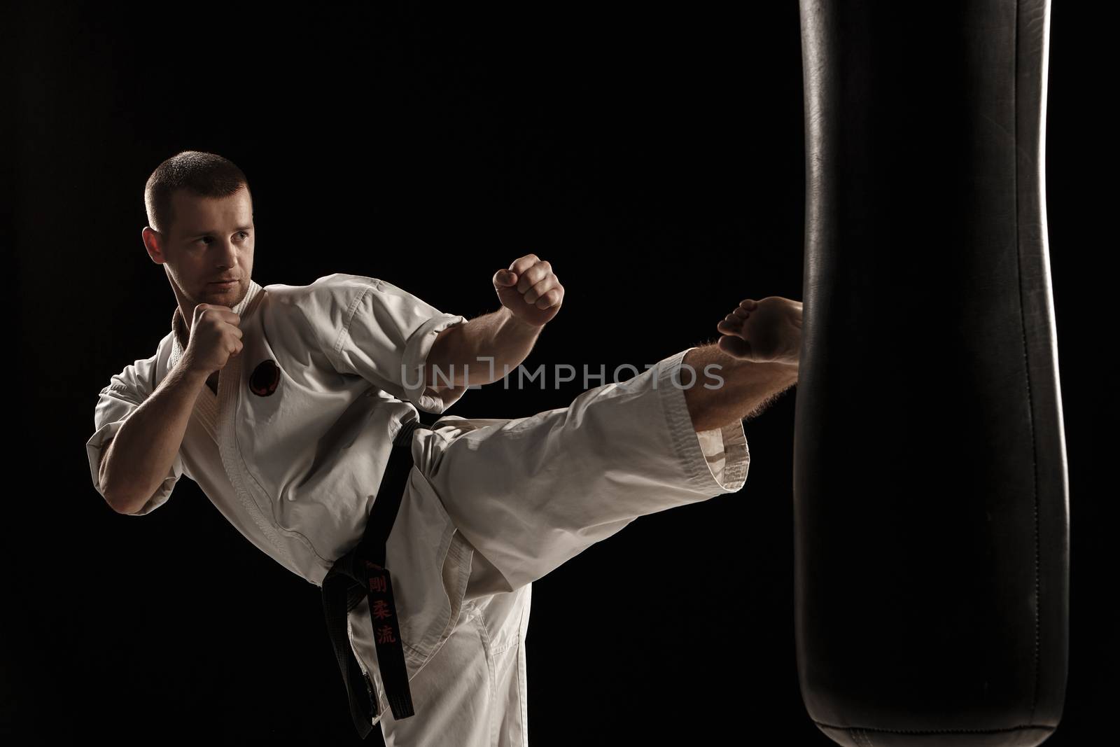 Man in white kimono training karate round kick in a punching bag over black background