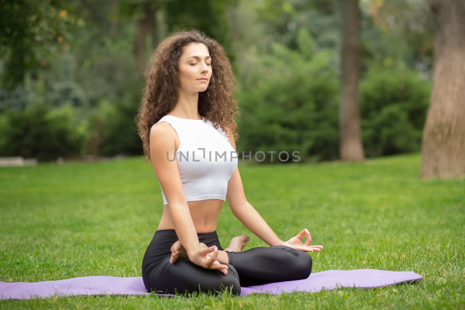 Pretty woman with eyes closed doing yoga meditation in the lotus position. Green grass background 