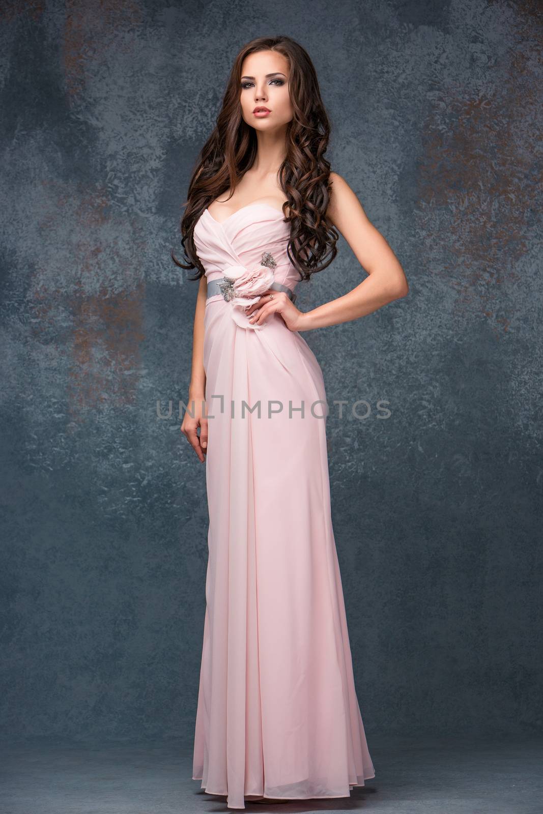 Sexy young brunette woman with her hair posing in full growth in a long pink dress in studio on gray background 