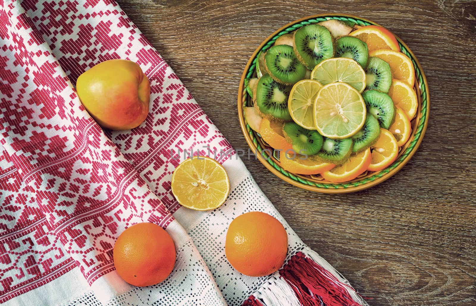 On the table is a dish of sliced fruits: oranges, kiwi, lemon. Near them on a beautiful towel are two oranges and an Apple.
