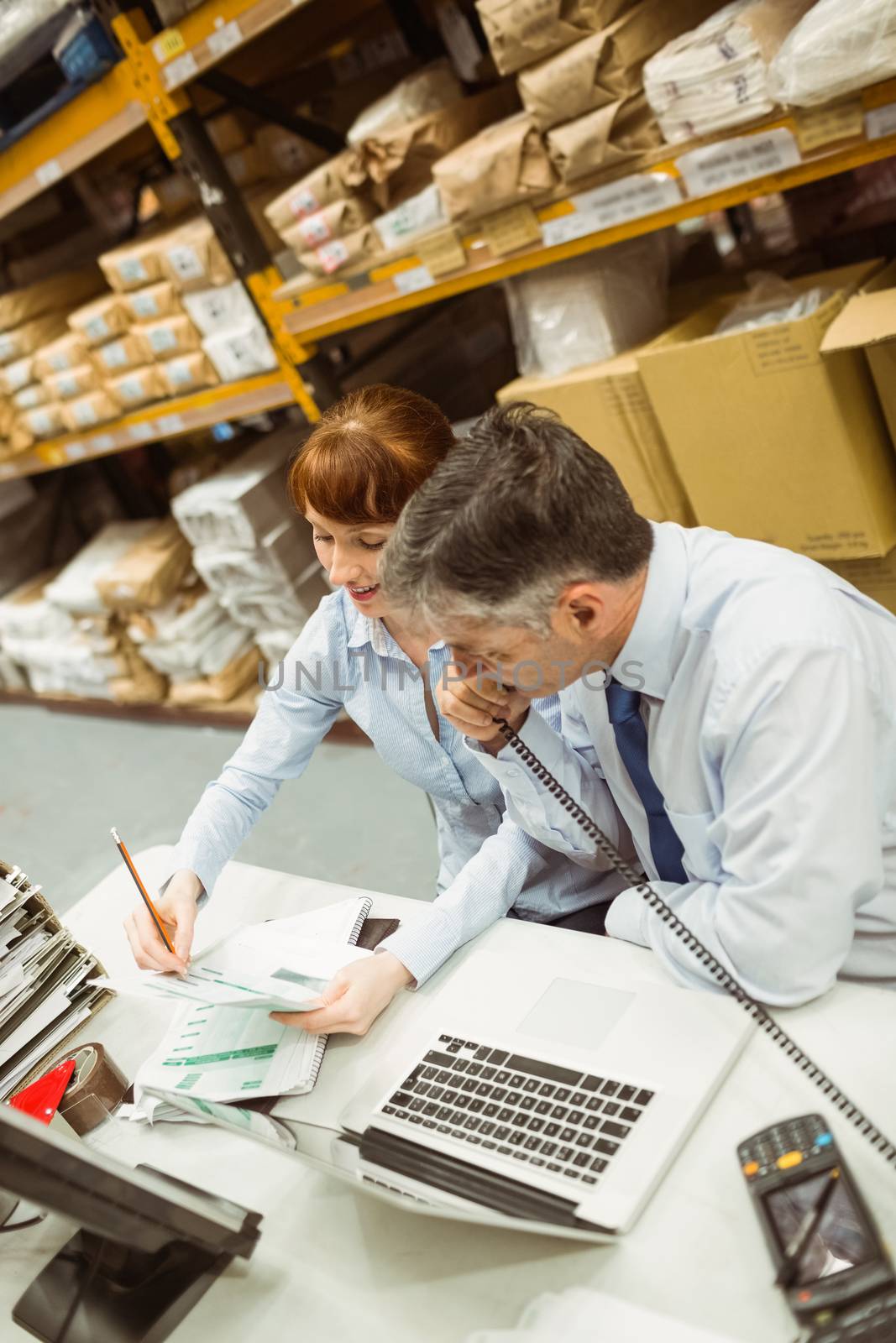 Manager working on laptop and talking on phone at desk in a large warehouse