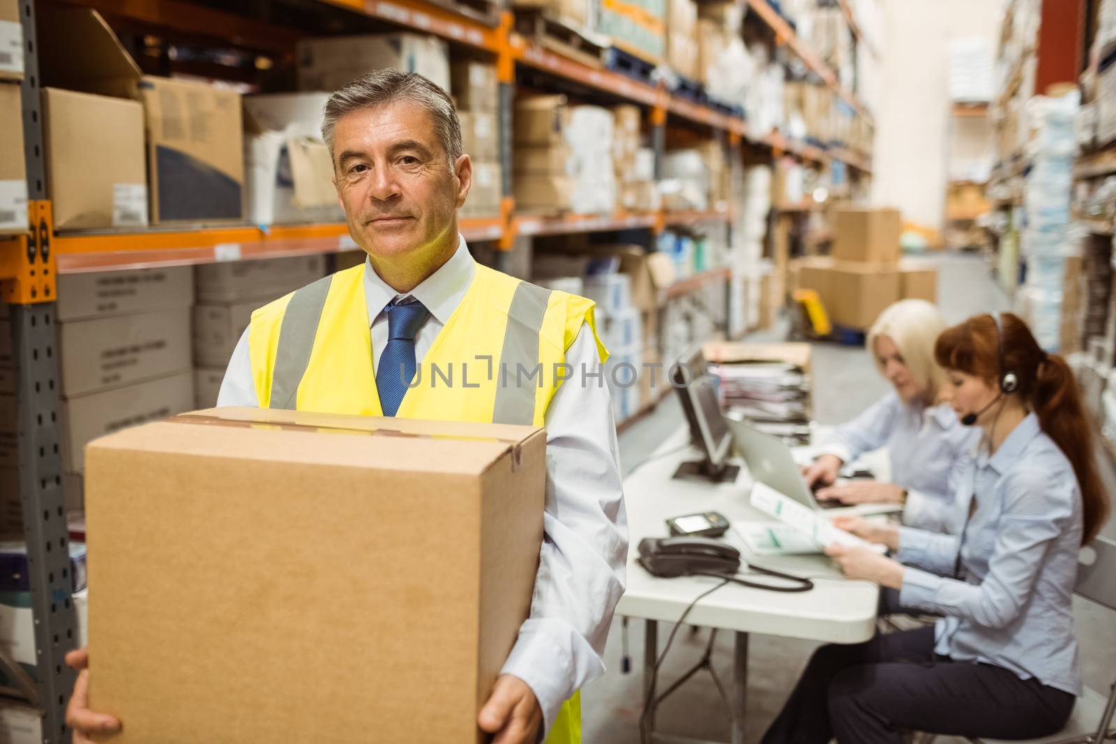 Warehouse manager smiling at camera carrying a box in a large warehouse