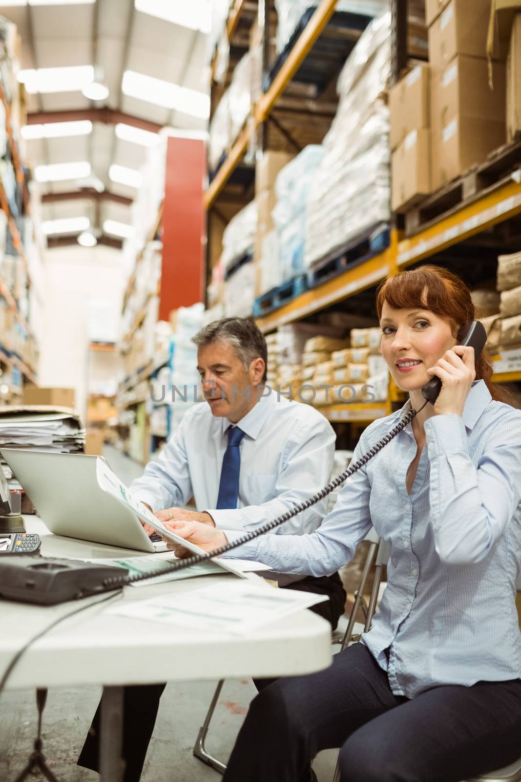 Manager working on laptop and talking on phone at desk in a large warehouse