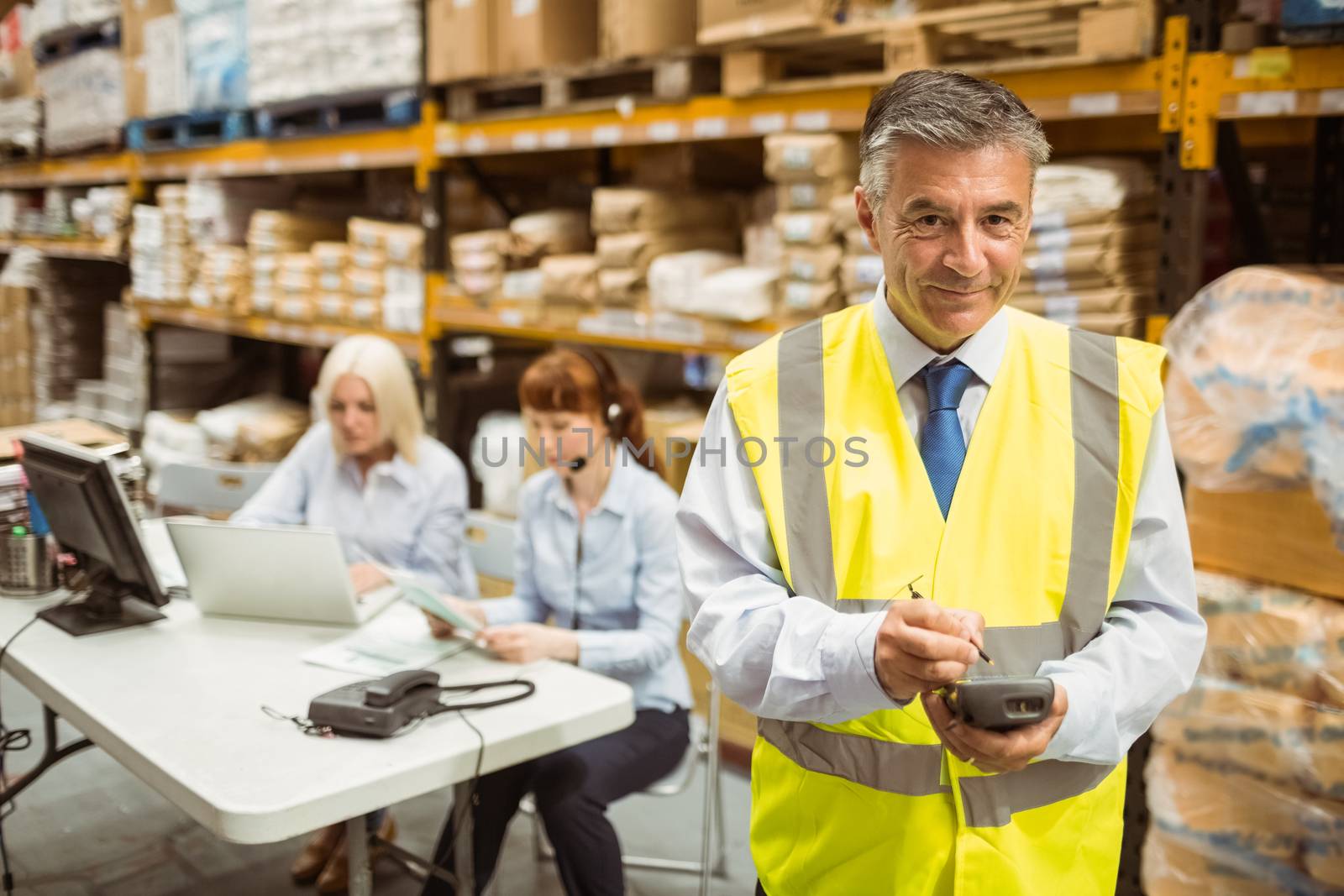 Smiling manager wearing yellow vest using handheld in a large warehouse