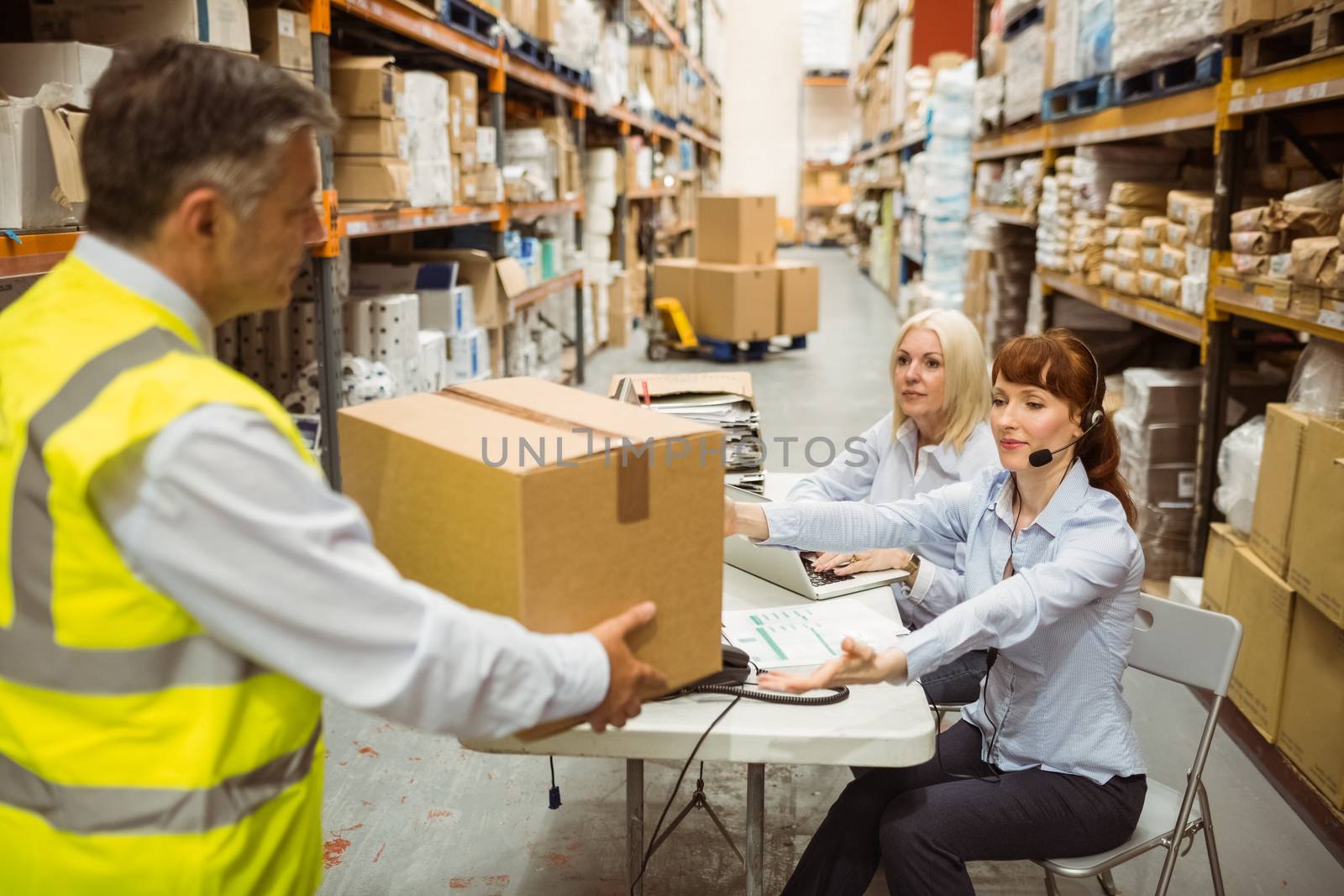 Manager wearing yellow vest giving box to his colleague in a large warehouse