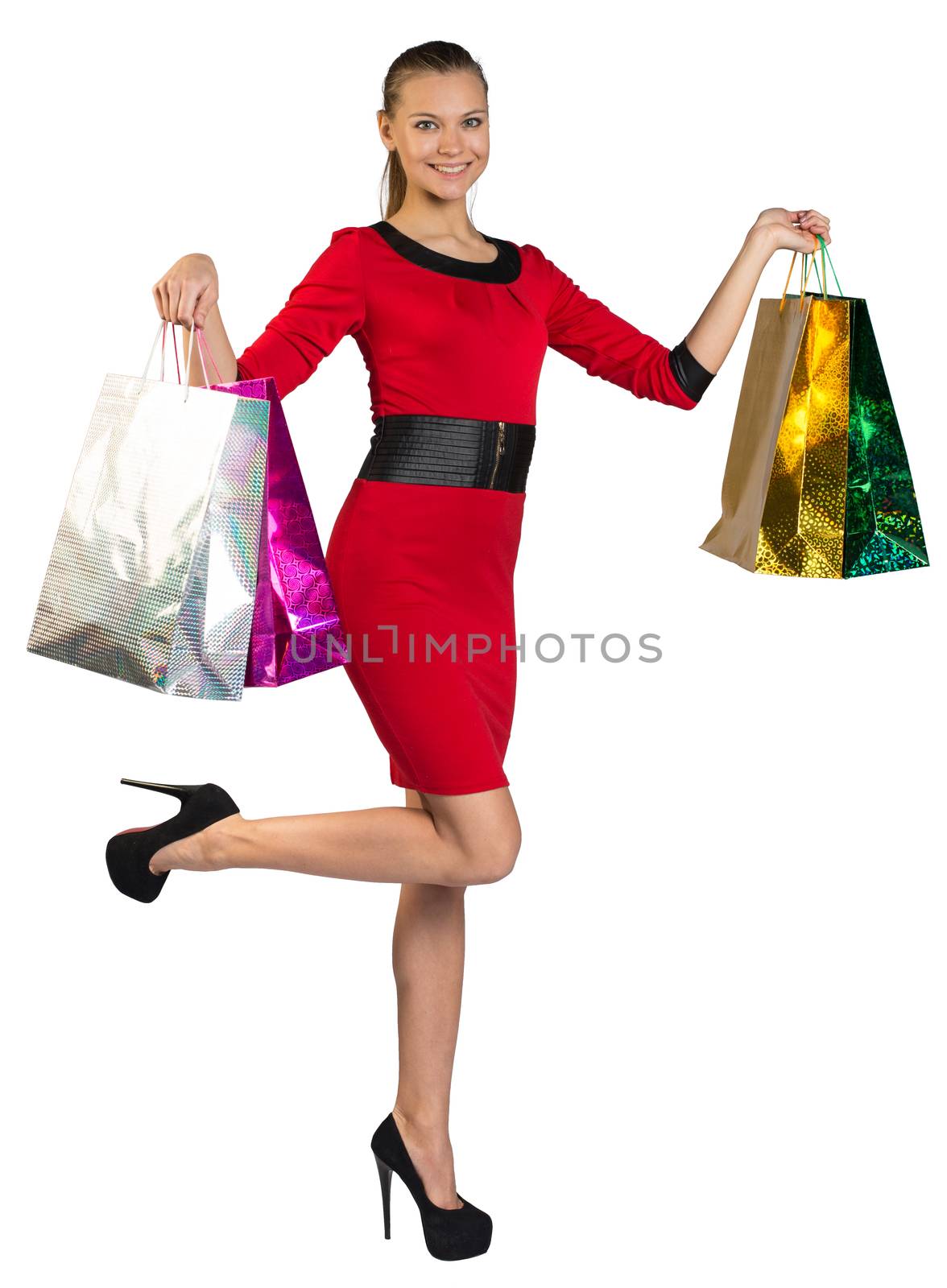 Half turned young woman with teeth smile and right leg up handing colorful shopping bags up and looking at camera. Isolated background