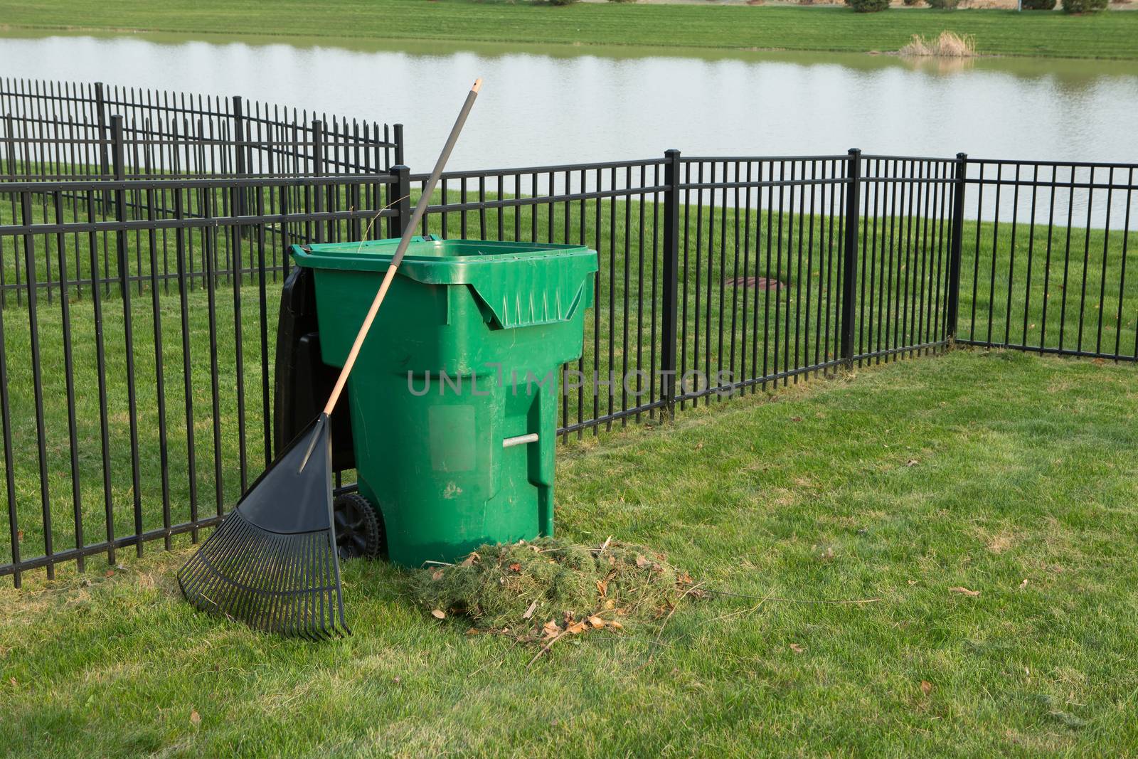 Yard maintenance in spring with a fresh heap of grass clippings and a rake leaning on a green plastic bin for composting organic waste on a neat lawn with wrought iron fence above a lake