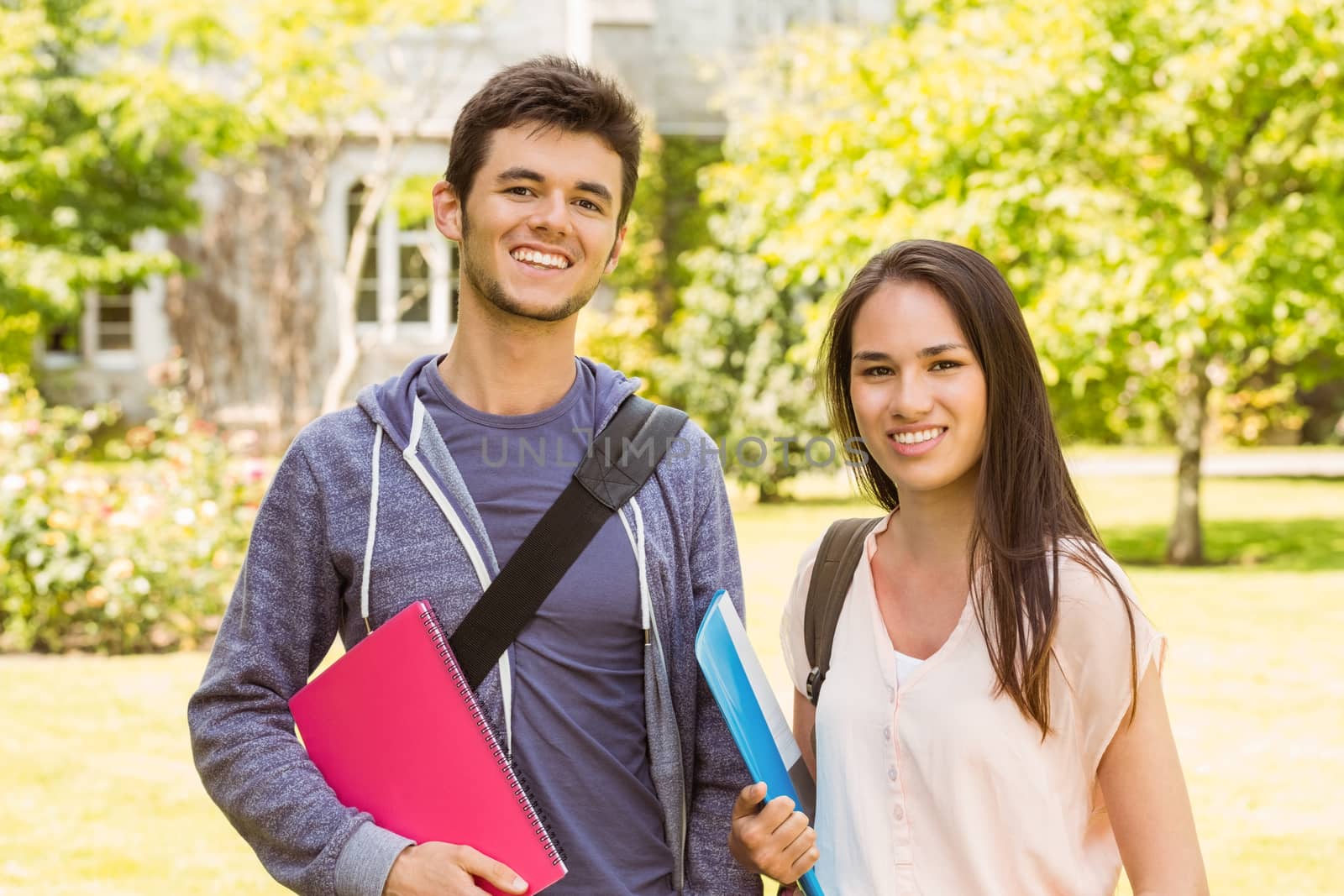 Smiling friends student standing with shoulder bag holding book in park at school