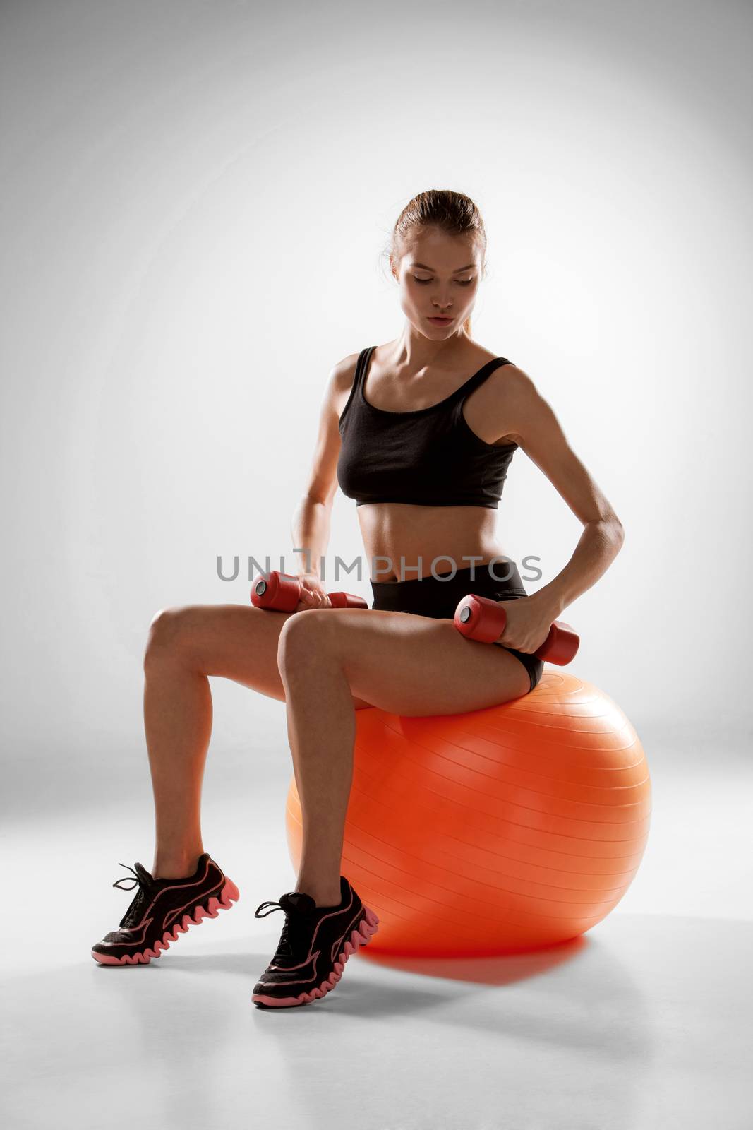 Sporty woman doing aerobic exercise with red dumbbells on a fitness ball on grey background