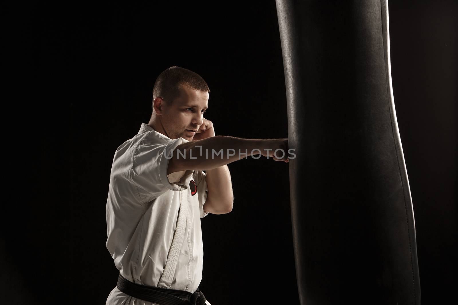 Man in white kimono training karate in a punching bag over black background