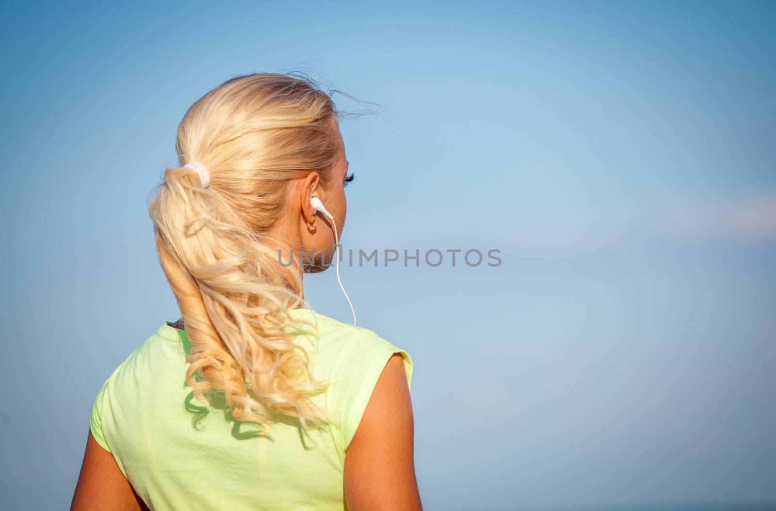 Attractive sporty young woman standing on the beach taking a break from exercising and listening to music with her mp4 player and head phones against a blue sky