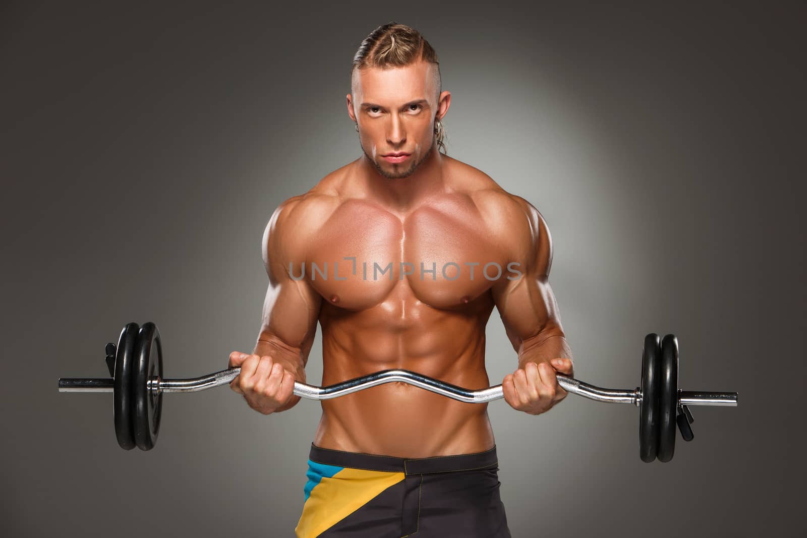 Portrait of super fit muscular young man working out in gym with barbell on gray background 