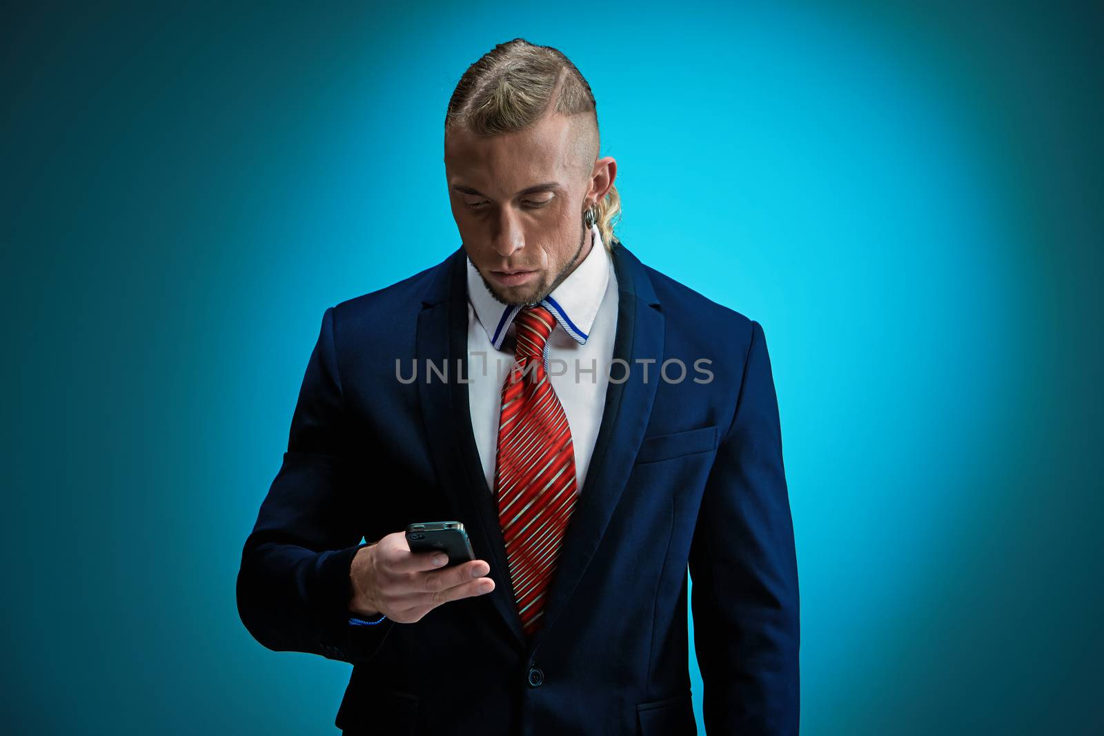 Portrait of an attractive businessman with phone wearing black suit. Blonde hair. On blue background