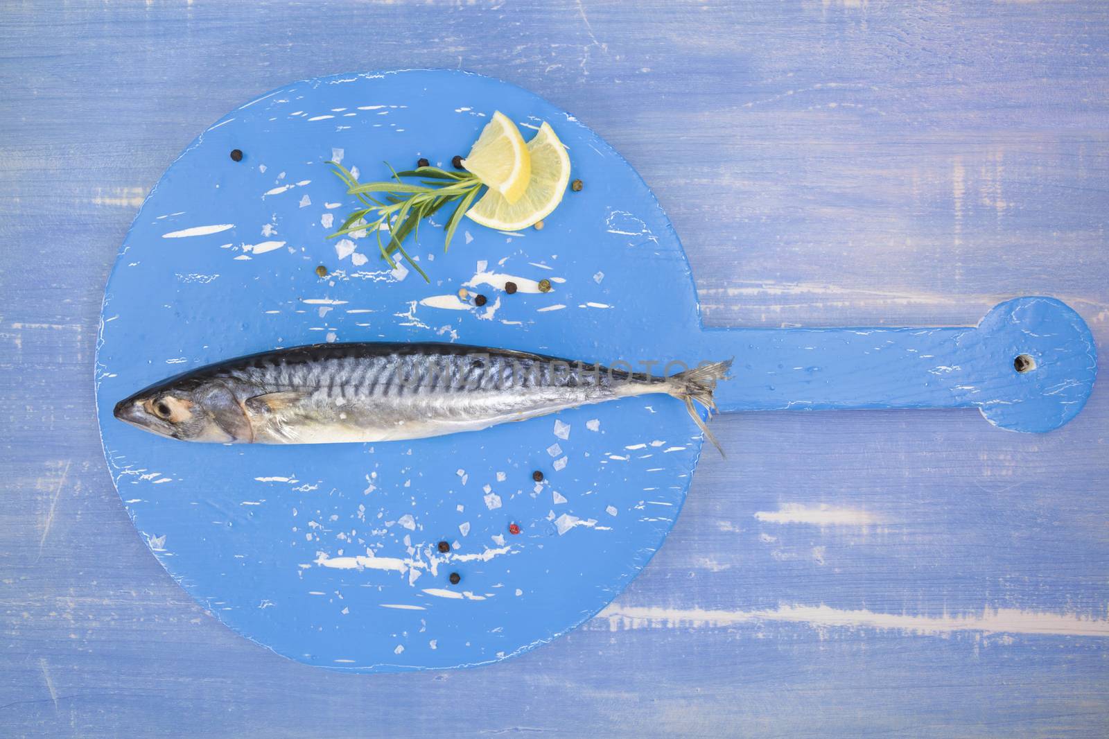 Delicious fresh mackerel fish on wooden kitchen board with lemon, rosemary and salt flakes on blue textured wooden background. Culinary healthy cooking.