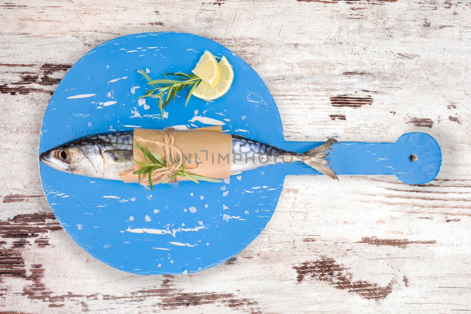 Delicious fresh mackerel fish on wooden kitchen board with lemon, rosemary and salt flakes on white textured wooden background. Culinary healthy cooking.