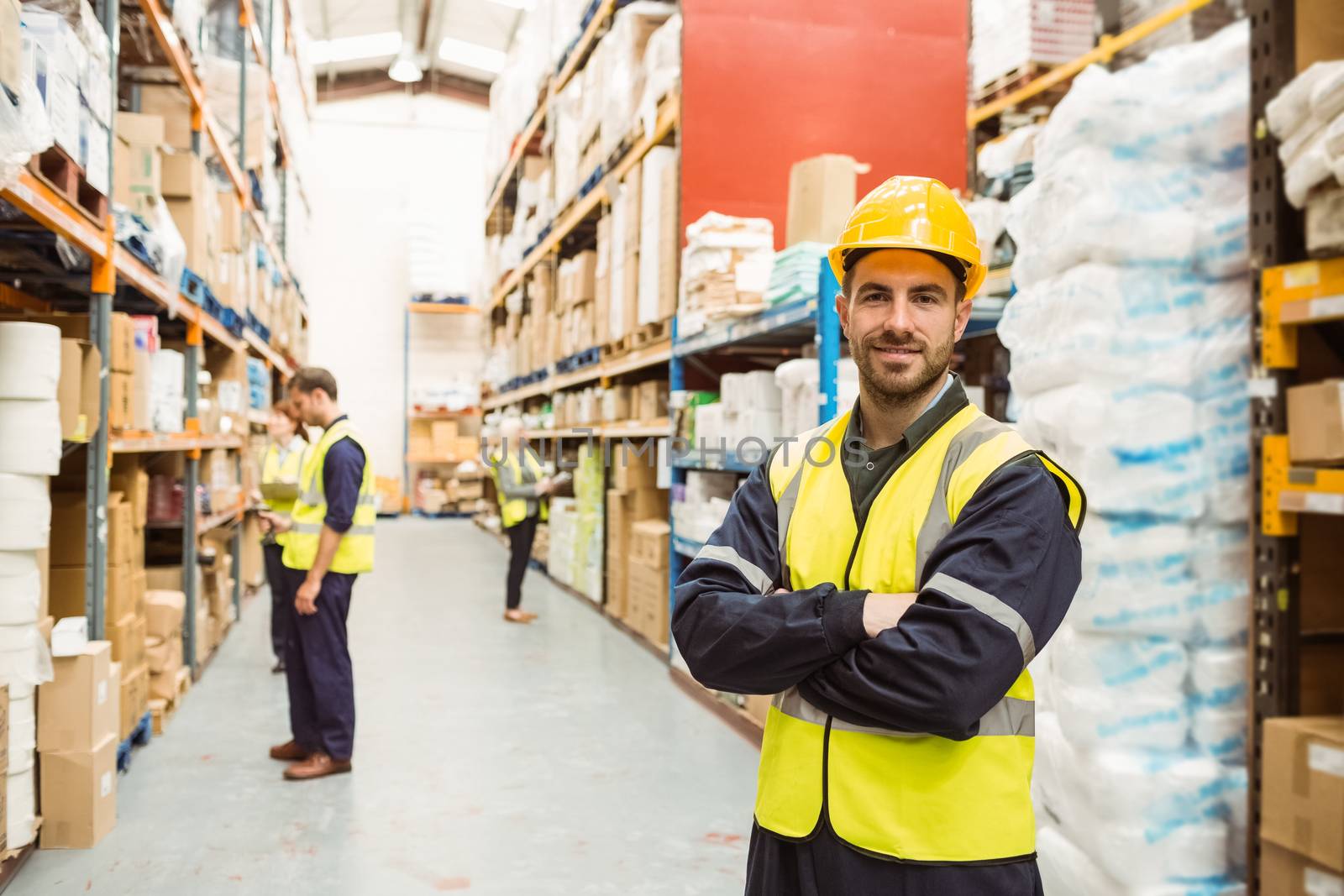 Smiling worker wearing yellow vest with arms crossed in a large warehouse