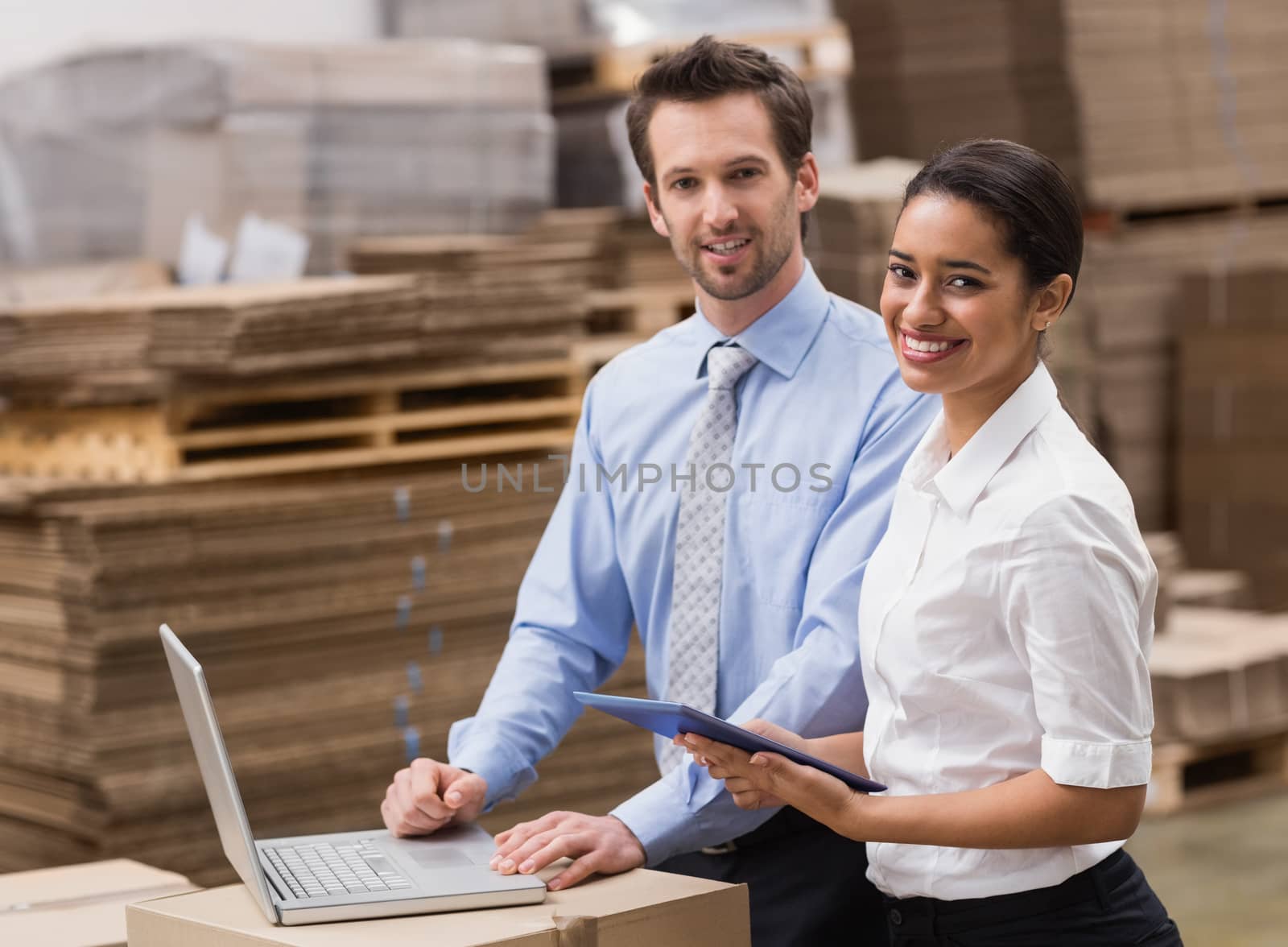 Managers working on laptop while smiling at camera in a large warehouse