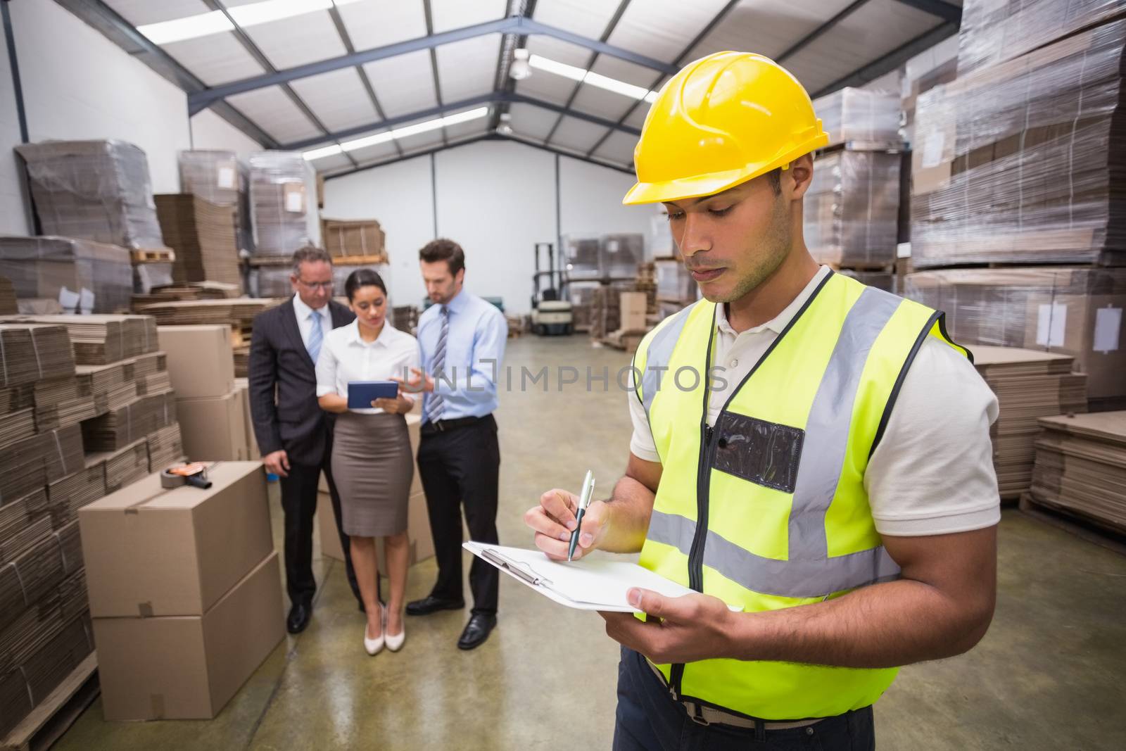 Worker writing on clipboard in front of his colleagues in a large warehouse