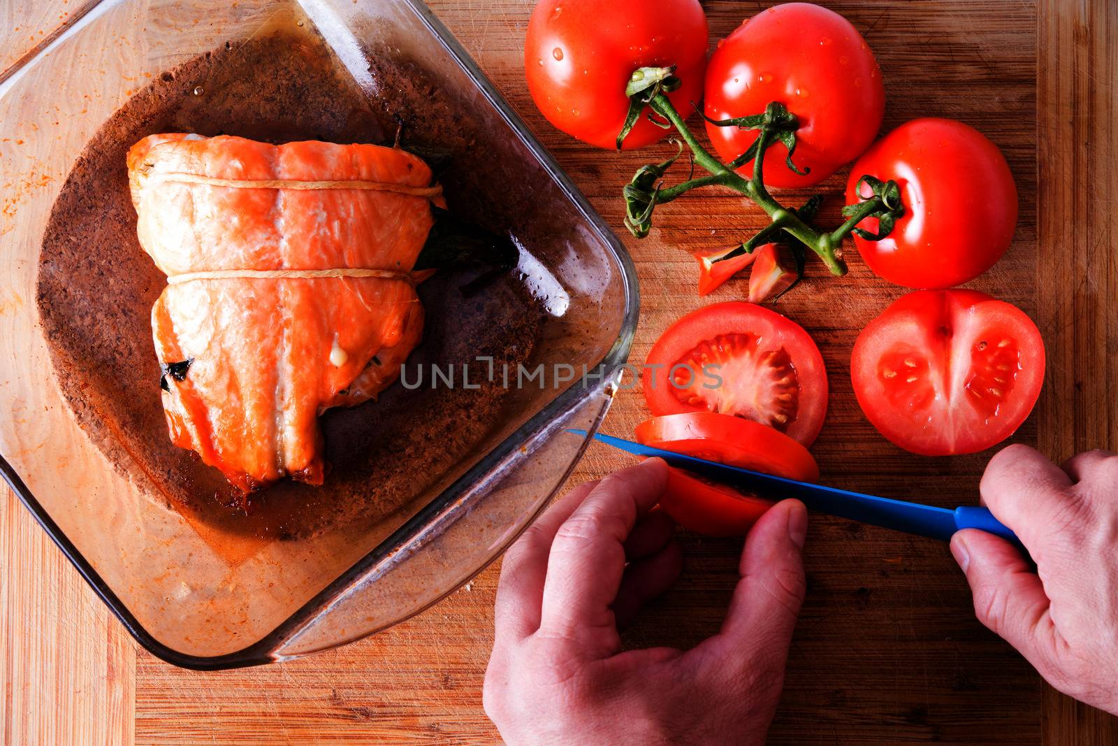 Chef preparing a gourmet salmon dinner slicing fresh vine tomatoes to accompany the succulent fish fillet in the glass oven dish alongside, view from above