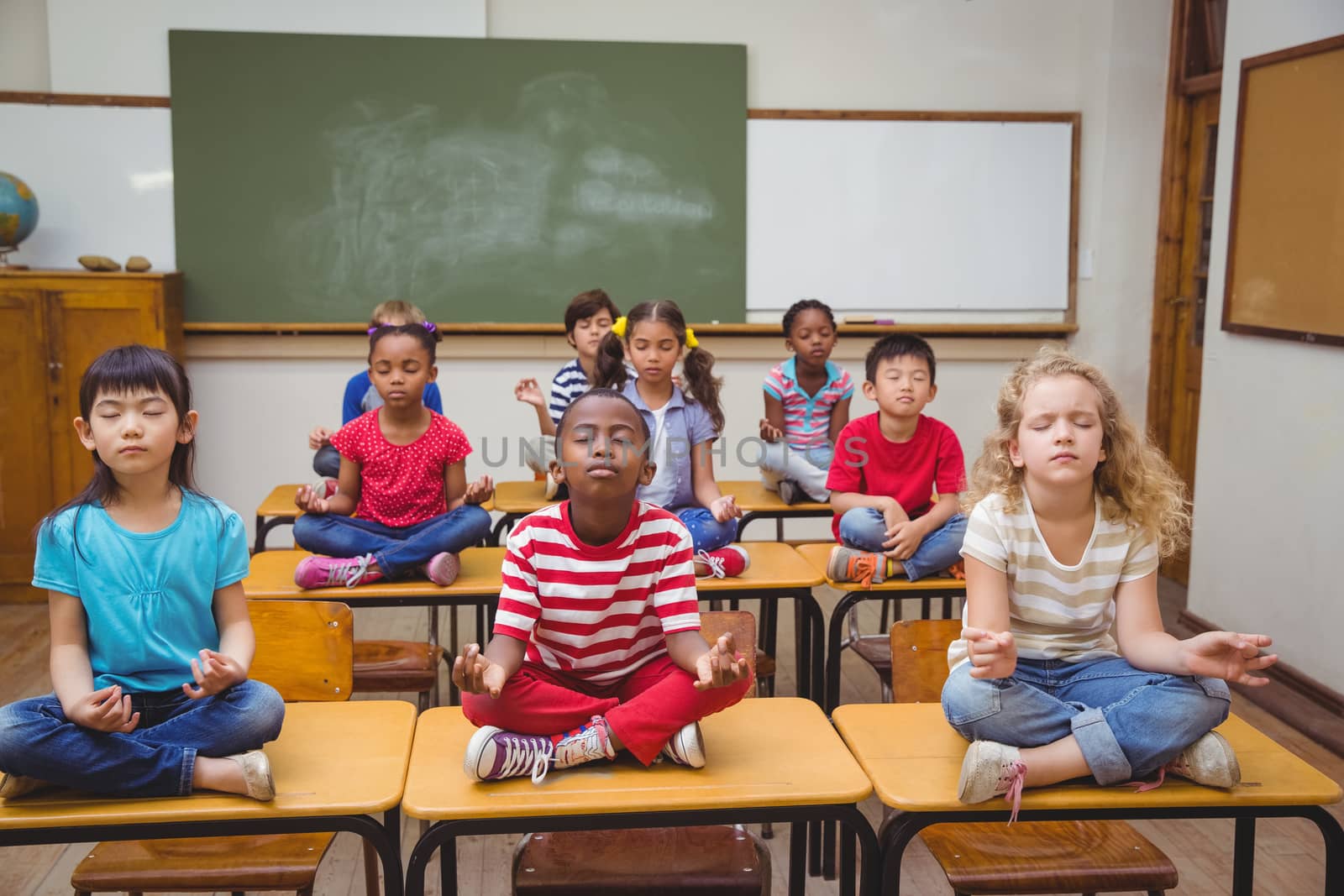 Pupils meditating in lotus position on desk in classroom by Wavebreakmedia