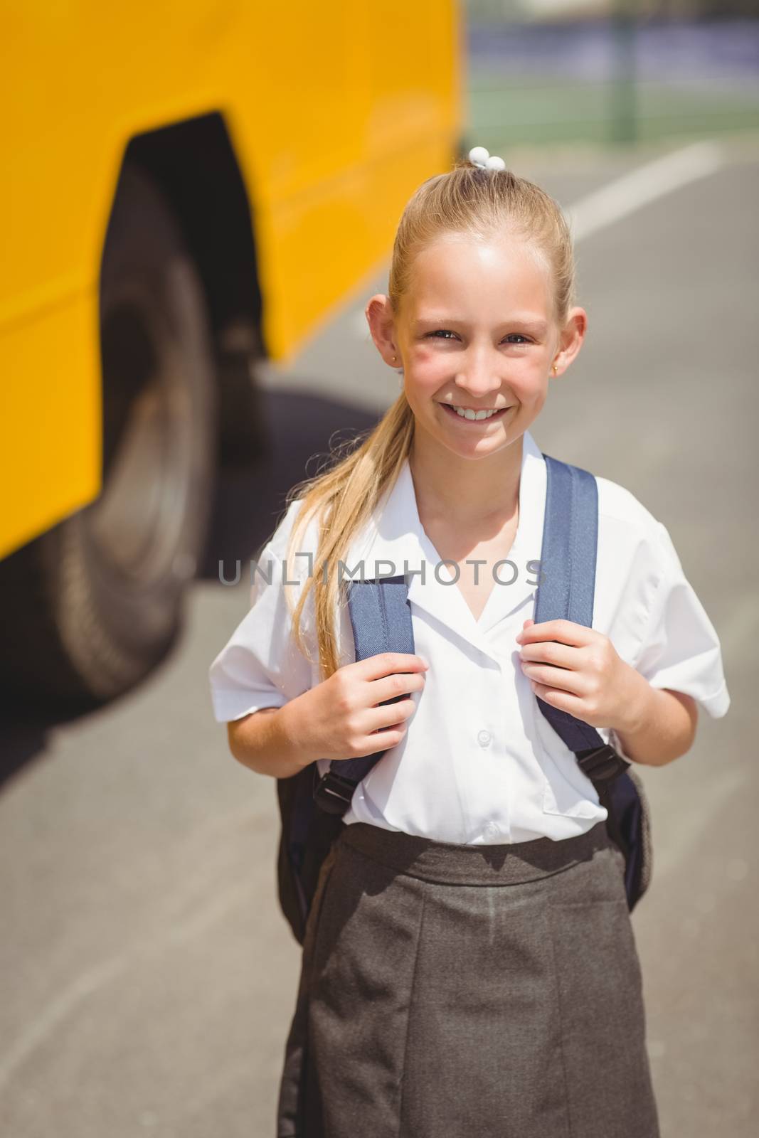 Cute pupil smiling at camera by the school bus outside the elementary school