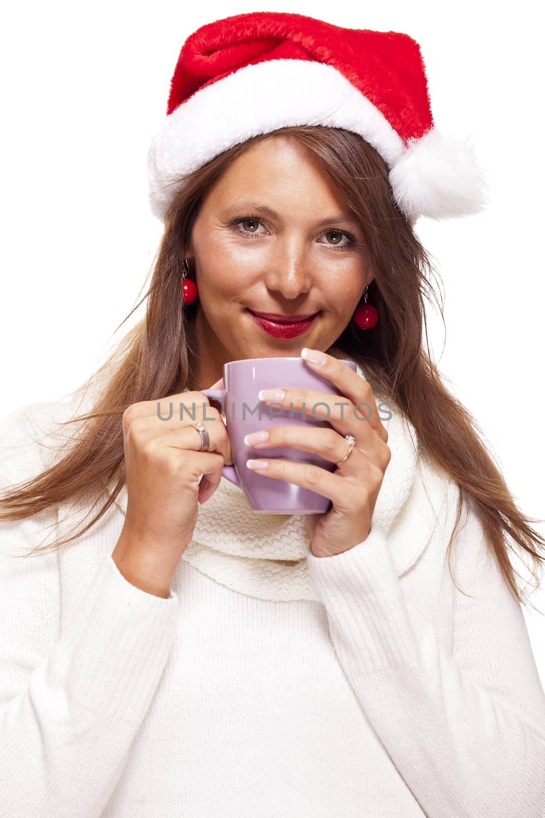 Cold attractive young woman with a cute smile in a festive red Santa hat sipping a hot mug of coffee that she is cradling in her hands to warm up in the winter weather, on white