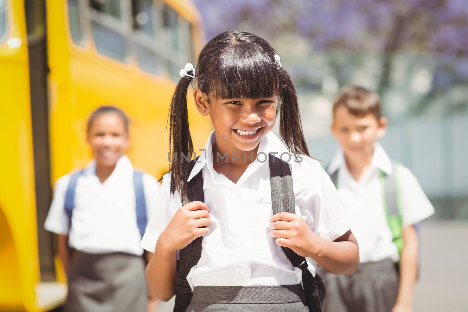 Cute pupil smiling at camera by the school bus outside the elementary school