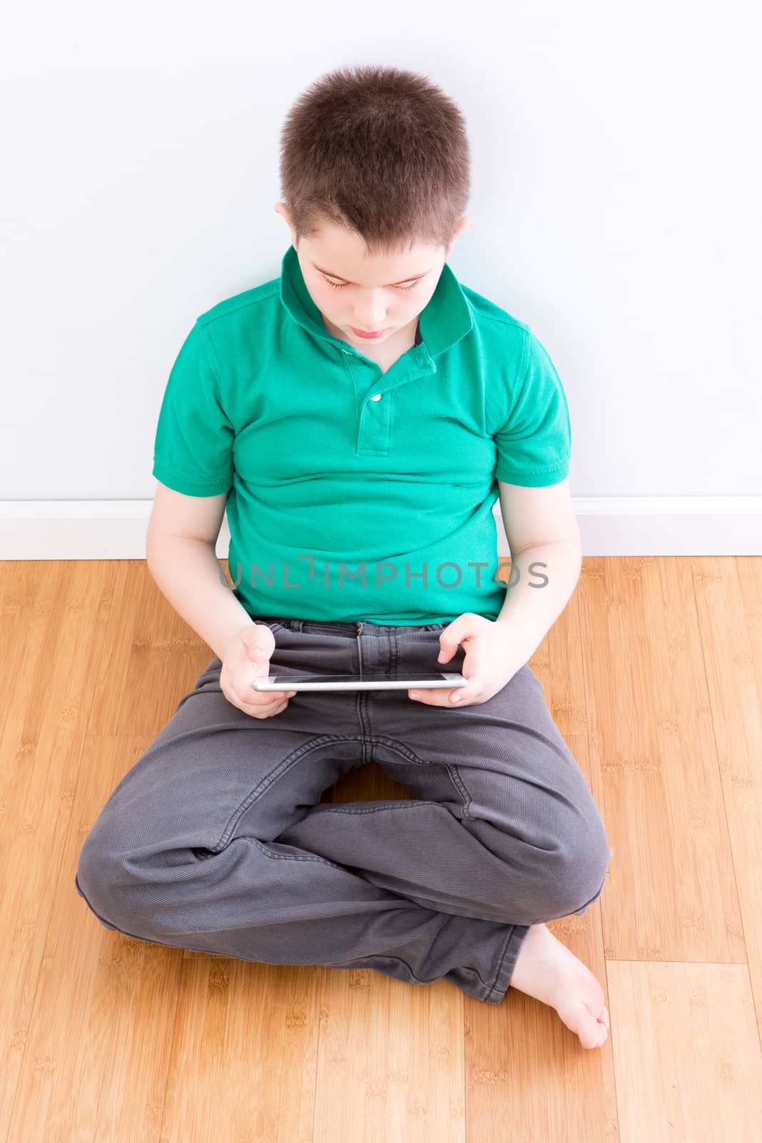 10 Year Old Young Boy Sitting on the Floor with Legs Crossed, Holding a Tablet Computer While Leaning on the Wall, Captured in High Angle View.