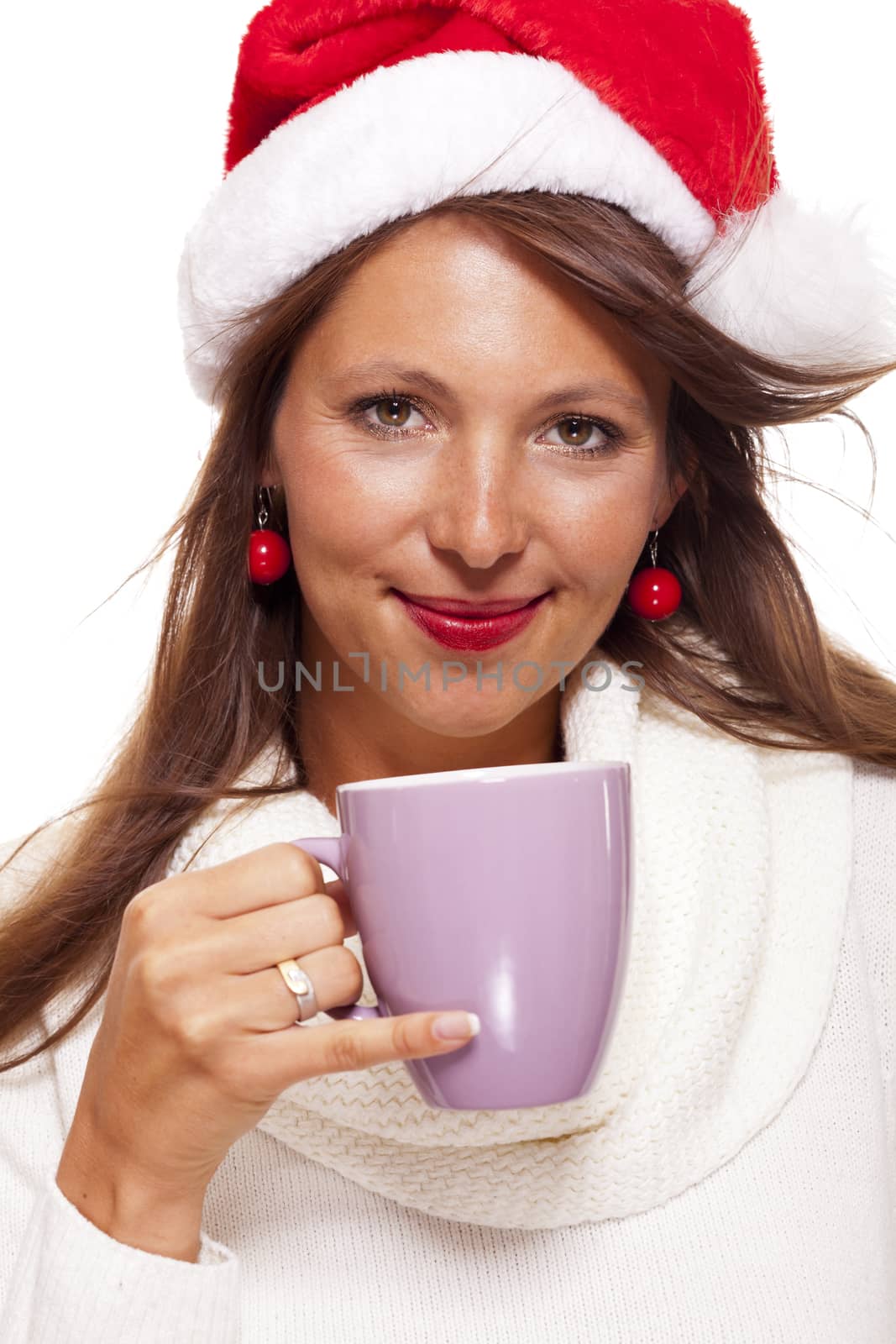 Cold attractive young woman with a cute smile in a festive red Santa hat sipping a hot mug of coffee that she is cradling in her hands to warm up in the winter weather, on white