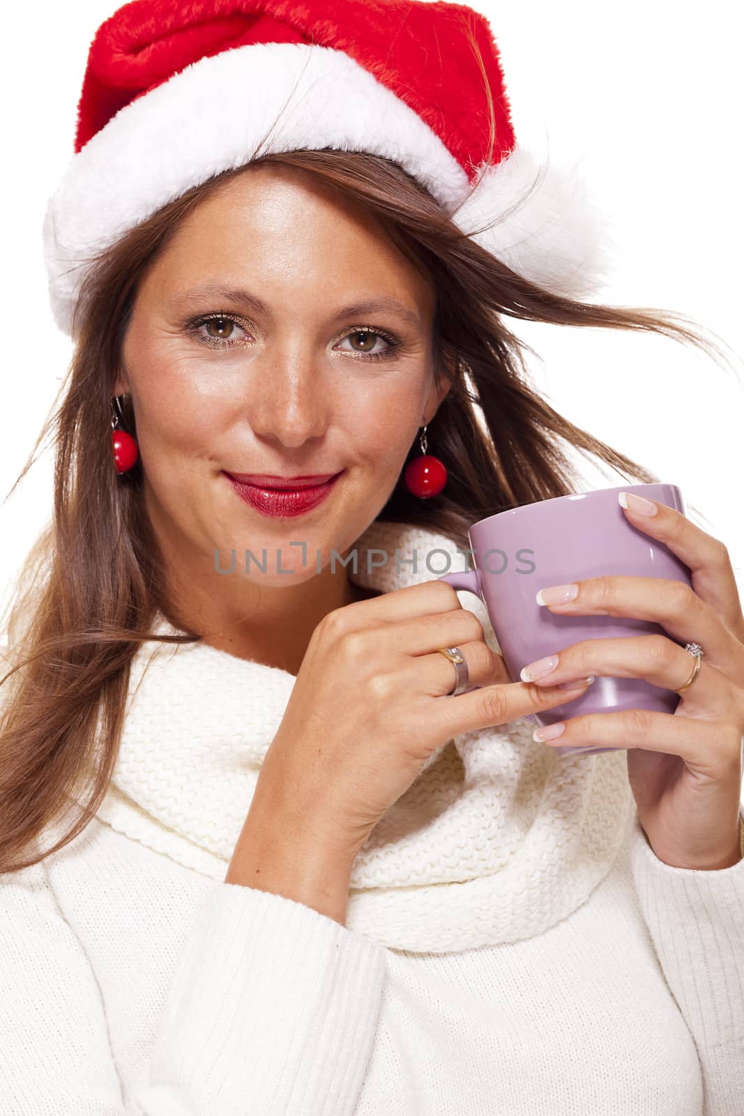 Cold attractive young woman with a cute smile in a festive red Santa hat sipping a hot mug of coffee that she is cradling in her hands to warm up in the winter weather, on white