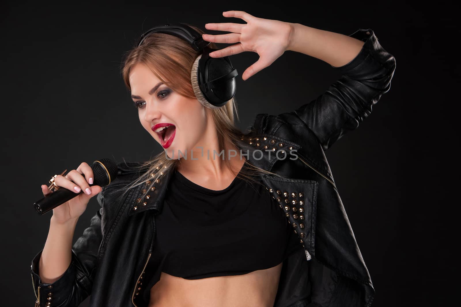 Portrait of a beautiful blonde young woman singing into microphone with headphones in studio on black background