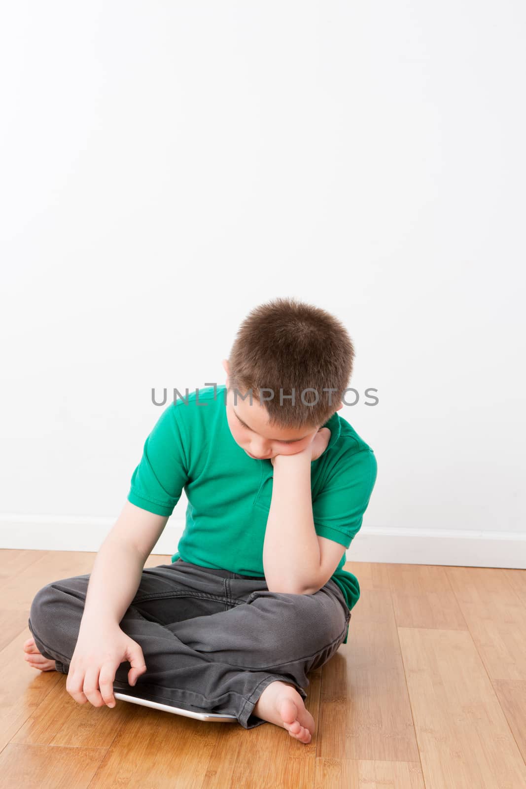 Serious Young Boy Sitting on the Floor and Leaning on his Hand While Browsing at his Tablet Computer.