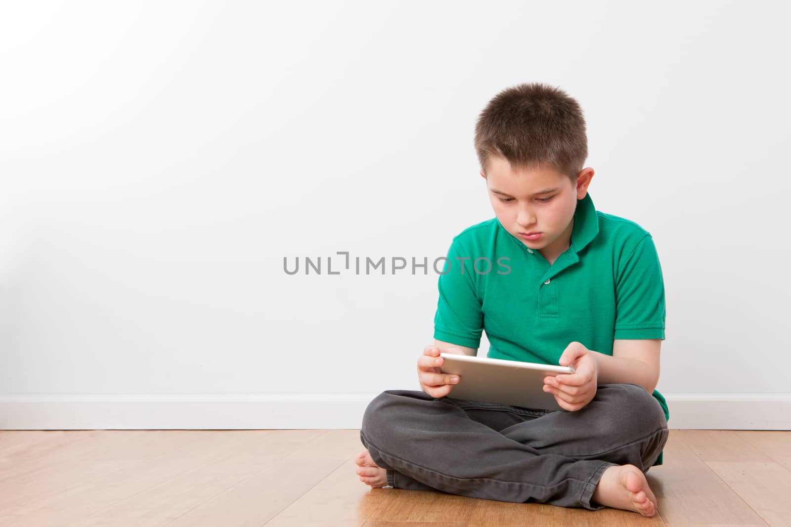 Serious Cute 10 Year Old Young Boy Sitting on the Floor with Legs Crossed, Busy with his Tablet Computer