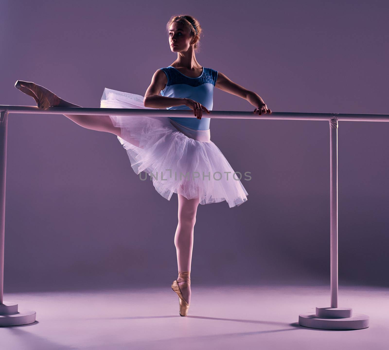 classic ballet dancer in white tutu posing on one leg at ballet barre on a lilac background