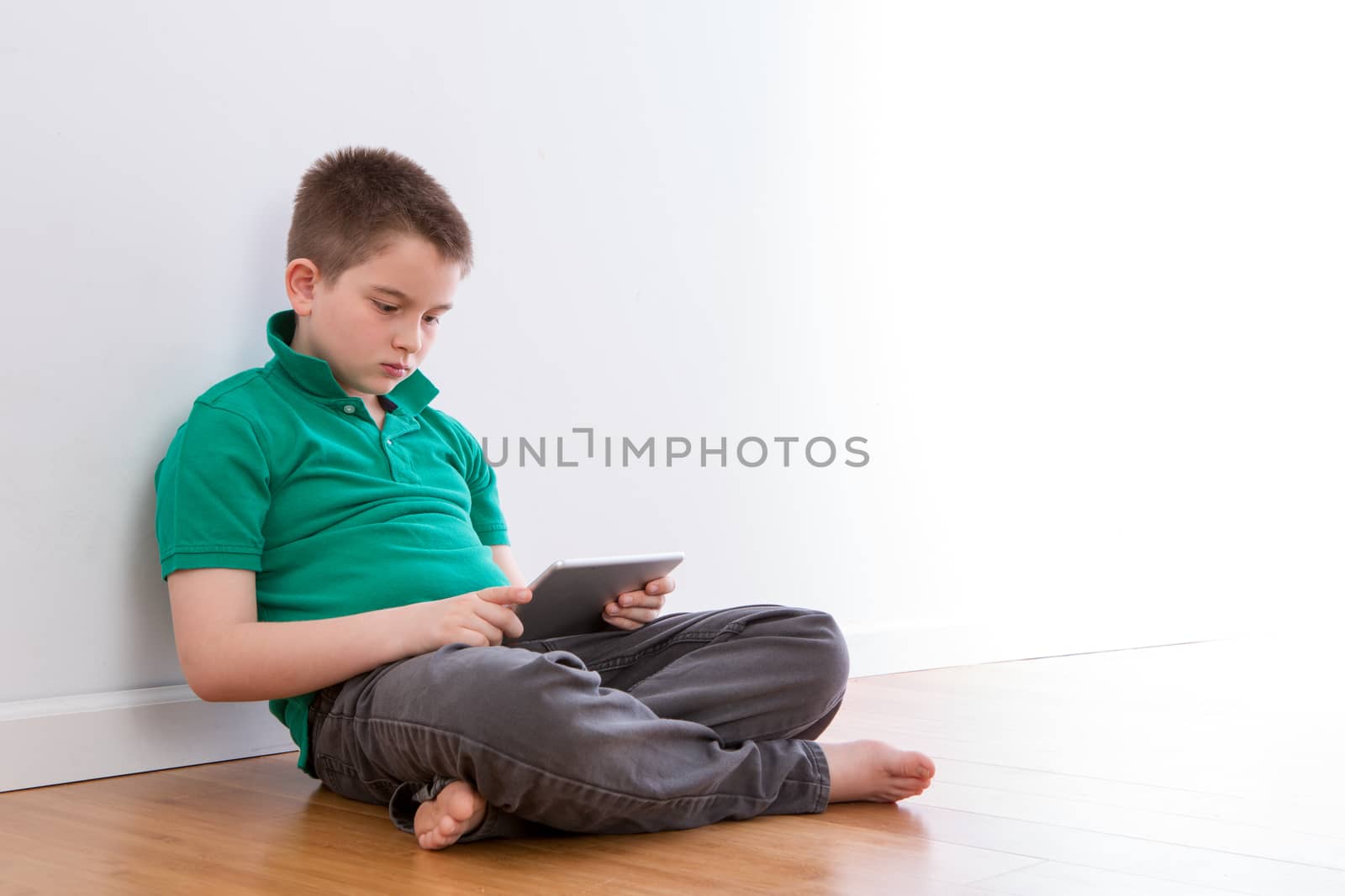 Serious 10 Year Old Male Kid Sitting on the Floor, Browsing at his Tablet Computer While Leaning on the White Wall.