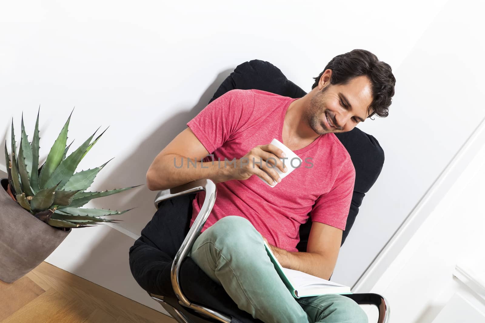 Young Man in Casual Clothing Sitting on Black Chair While Reading a Book and Holding a Glass of Drink.