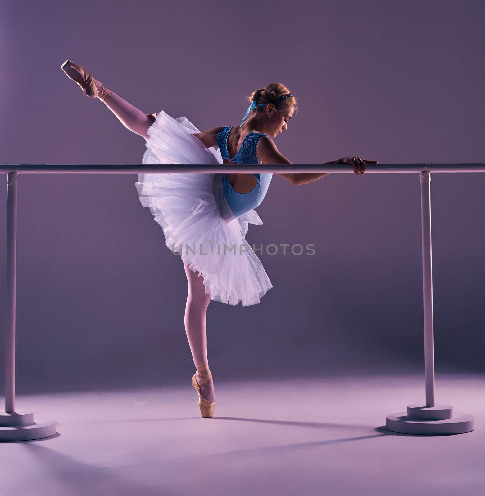 classic ballet dancer in white tutu posing on one leg at ballet barre on a lilac background