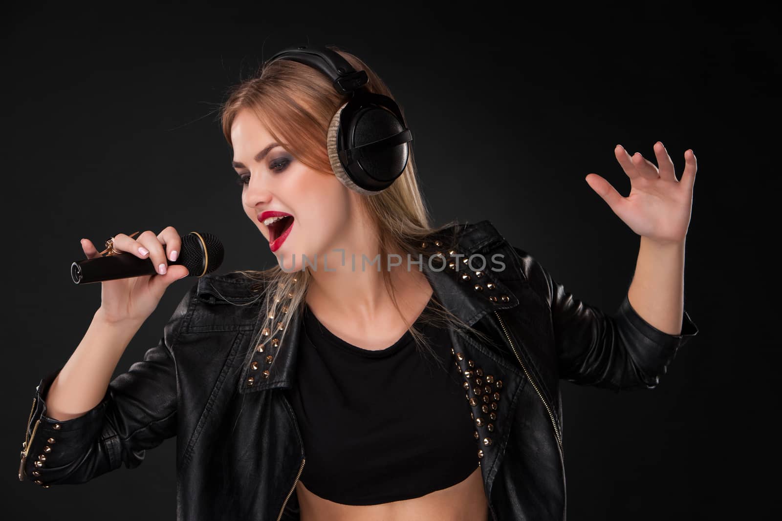 Portrait of a beautiful blonde young woman singing into microphone with headphones in studio on black background