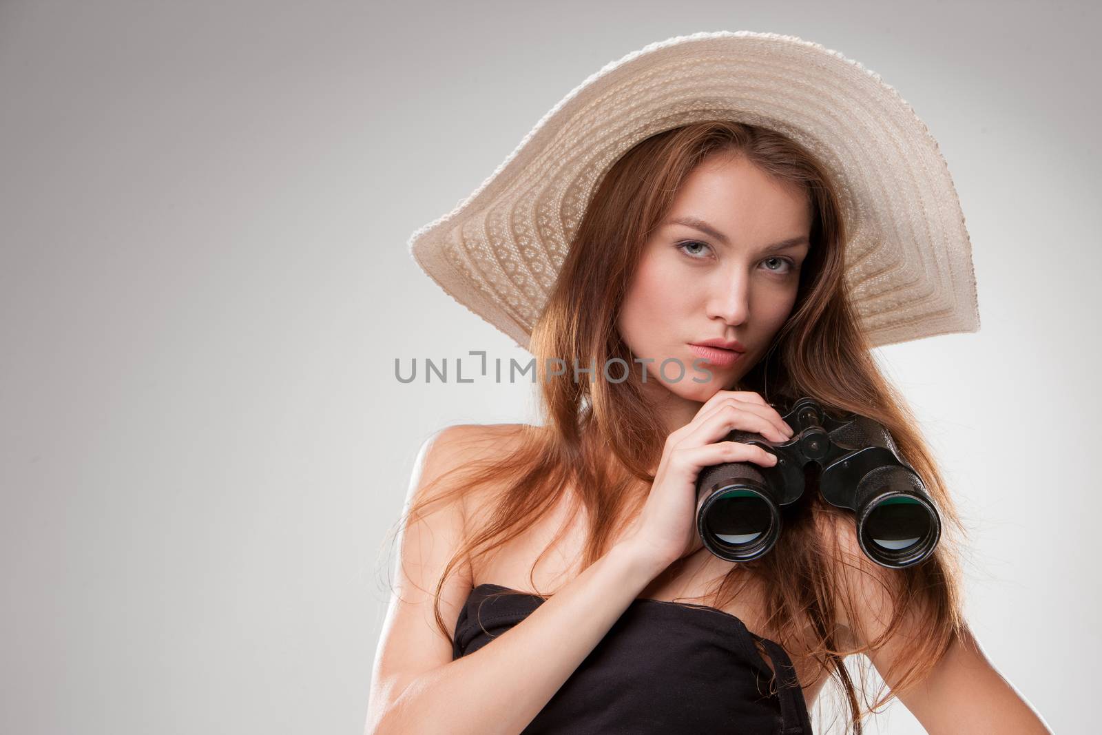 Young woman in hat with binoculars isolated on gray background. Travel and adventure concept. Closeup.