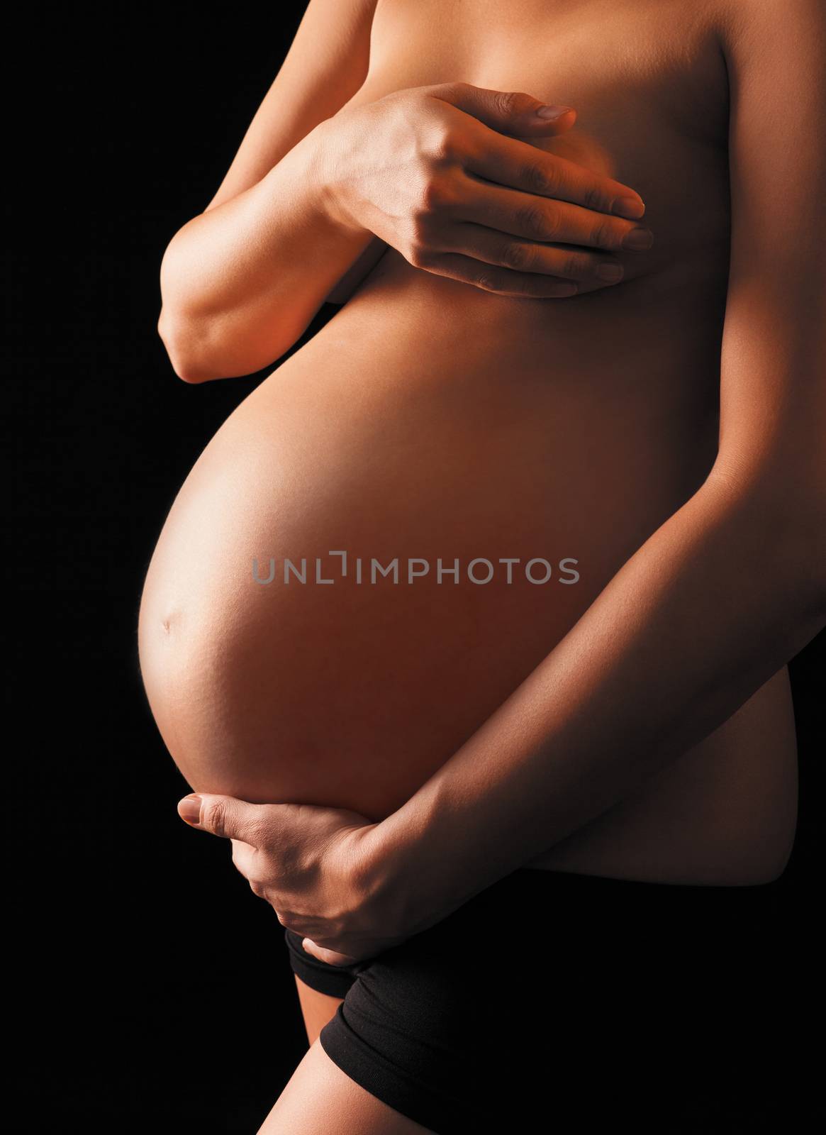 Pregnant topless woman hiding her breasts is holding her abdomen on black background, isolated with work path.