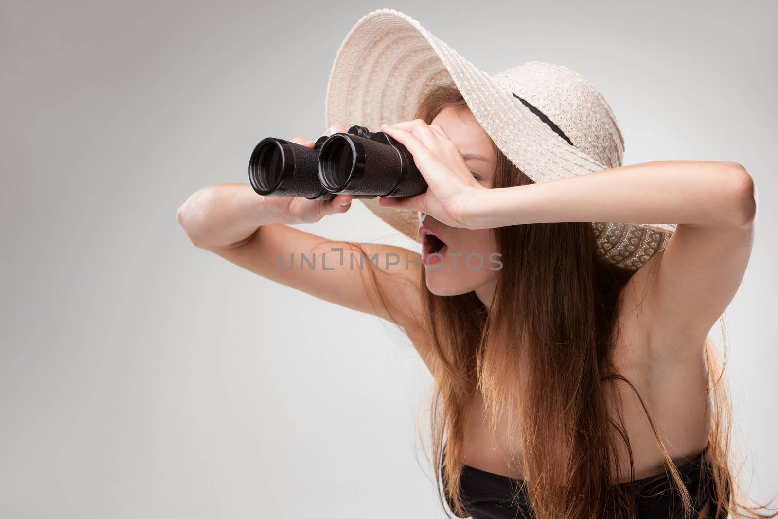 Young woman in hat looking through binoculars on gray background. Travel and adventure concept. Closeup.