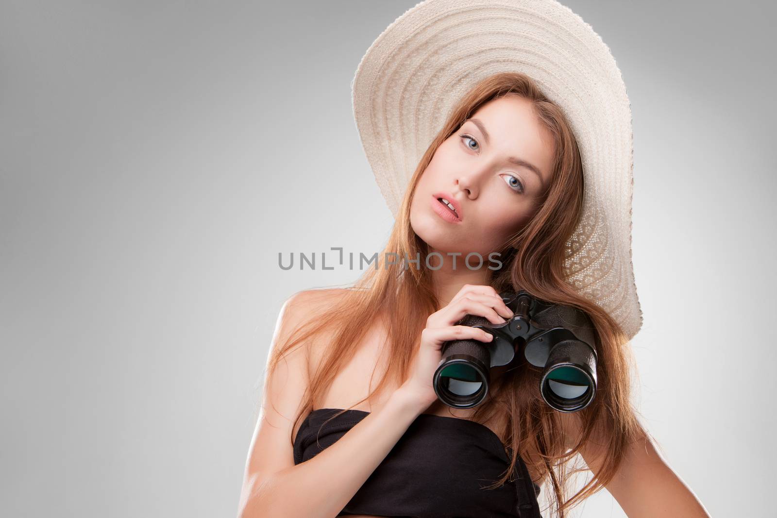 Young woman in hat with binoculars isolated on gray background. Travel and adventure concept. Closeup.