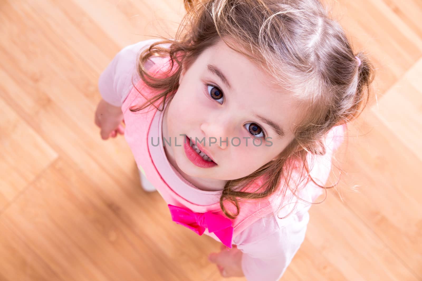 Pretty little girl with curly hair and beseeching eyes standing looking up into the camera as she asks for something that she wants, view looking down into her face with wood floor background