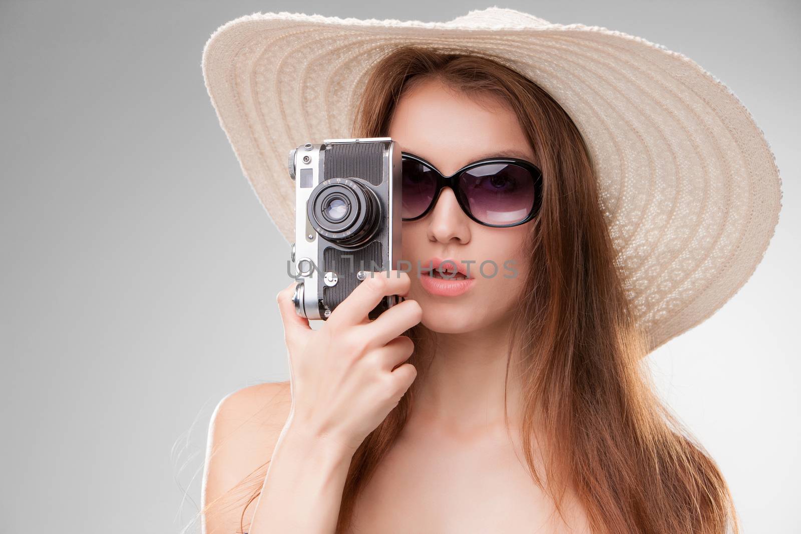 Girl in broad-brimmed hat and sunglasses with retro camera isolated on gray background