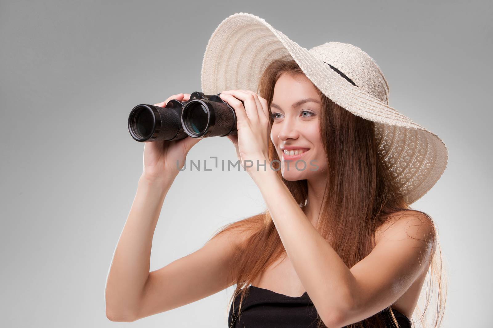 Young woman in hat looking through binoculars isolated on gray background. Travel and adventure concept. Closeup.