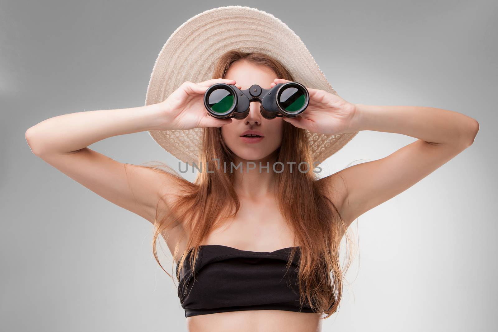 Young woman in hat with binoculars isolated on gray background. Travel and adventure concept. Closeup.