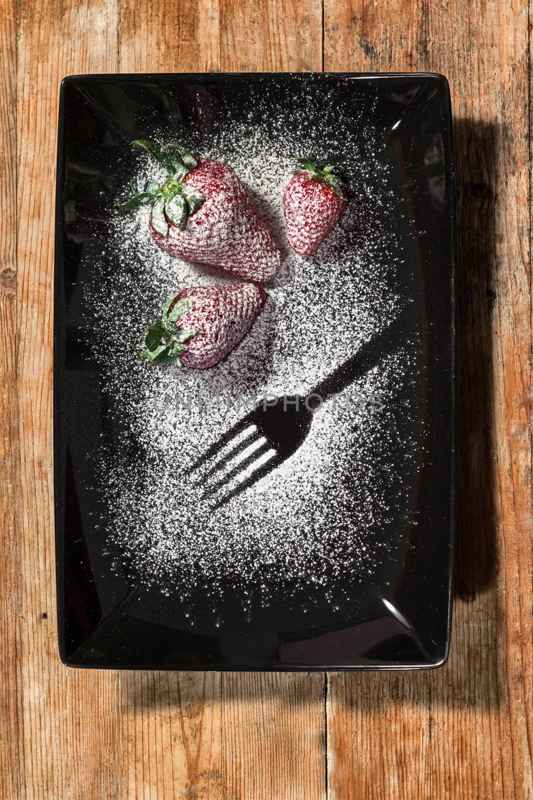three strawberries sprinkled with sugar on a black plate and wooden base