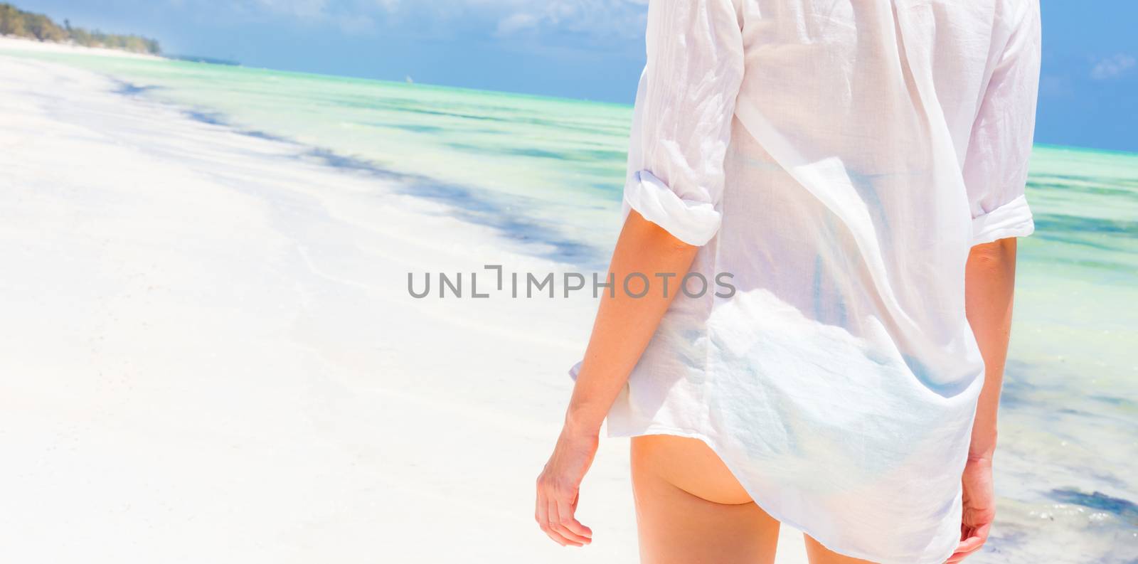 Happy woman enjoying, relaxing joyfully in summer by tropical blue water. Beautiful caucasian model  wearing white beach tunic on vacations looking down picture perfect Paje beach, Zanzibar, Tanzania.