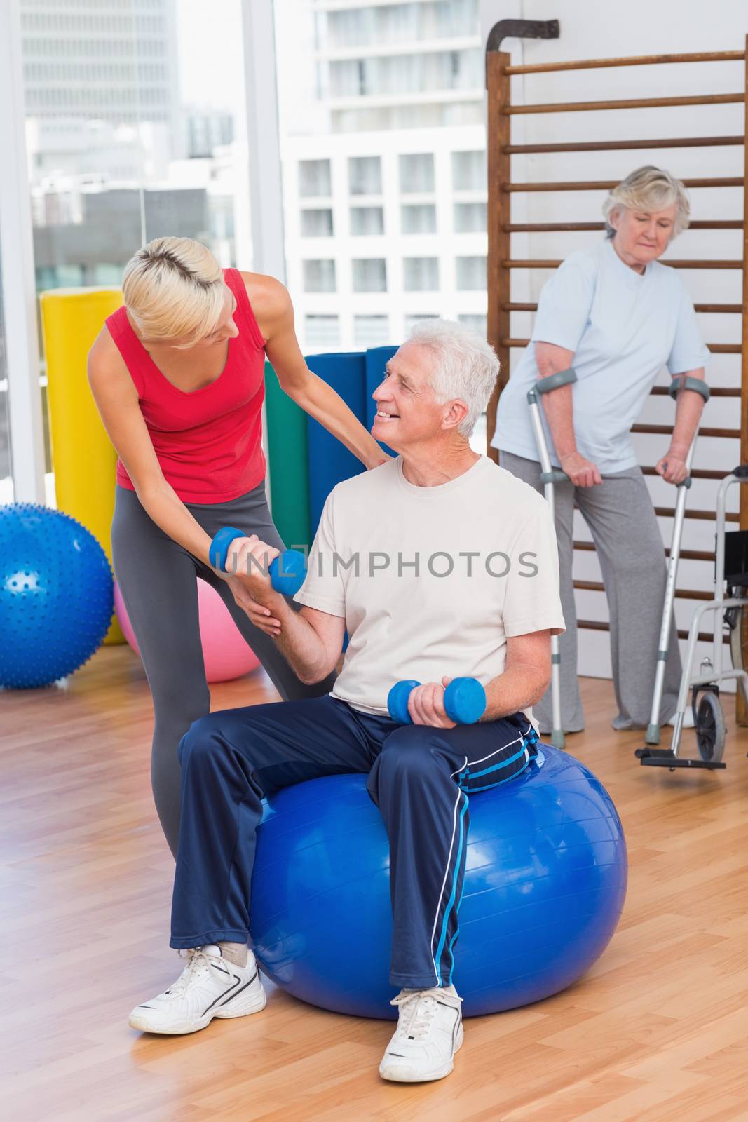Trainer assisting senior man in lifting dumbbells while woman using crutches in background at gym
